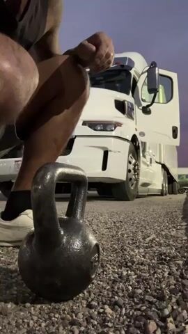 A man is performing kettlebell squats in front of a white semi-truck. The man is wearing a black Adidas tank top, gray shorts, and white sneakers. He has tattoos on his arms. The semi-truck has its driver's side door open. The scene is outdoors on a gravel surface, with a purple and orange sky suggesting it is dusk or dawn. The truck has a license plate that reads "BLDQBB". There is some green foliage visible in the background on the right side of the frame. The man appears focused and determined as he executes the exercise.