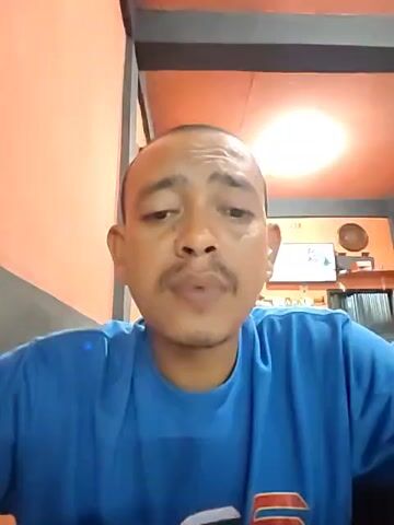 A man with a shaved head and mustache, wearing a blue t-shirt, is recording a video of himself. He is indoors, with an orange-colored wall in the background. There's a doorway or opening behind him with dark metal framing. A television screen is visible in the background, showing a nature scene. The lighting is bright. The man looks directly at the camera, speaks, and exhales smoke.