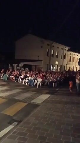 A nighttime outdoor crowd watches an unseen event. People are seated in rows of white plastic chairs arranged on a paved area, possibly a street, with a pedestrian crossing visible in the foreground. The backdrop consists of a building with lit windows. The audience appears engaged, with some individuals clapping. The lighting is dim, suggesting the event is happening after dark.