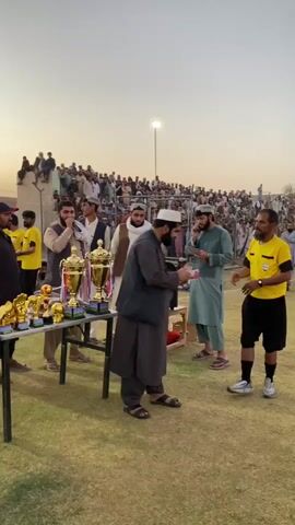 This is a vertical, medium shot of an outdoor award ceremony, likely a sporting event, with a crowd of spectators in the background. The sky is a pale orange, suggesting it is either sunrise or sunset. In the foreground, a table is laden with trophies, some of which are gold and appear to be of various sizes. Two men are in the center of the frame, shaking hands. The man on the left is wearing traditional clothing, including a dark robe and a white turban. The man on the right is dressed as a referee, wearing a bright yellow shirt with black trim, black shorts, black socks, and athletic shoes. He is also wearing a whistle around his neck. The man on the left is handing a folded piece of paper, possibly a certificate or award, to the referee. Behind them, a crowd of men, many wearing turbans and traditional clothing, are gathered, some seated on bleachers and others standing. A lone light pole is visible in the distance. The ground is covered in green grass.