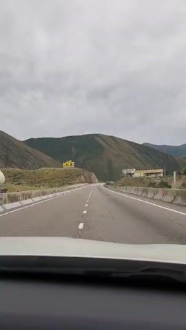 The video captures a view from inside a car driving on a paved road. The road is divided by white lines, indicating multiple lanes. On either side of the road are concrete barriers. In the distance, on a hill to the left, a large yellow sign with Cyrillic letters is visible. Further down the road, on the right, is a building complex situated on another hill. The landscape is mountainous, with green slopes and a cloudy sky overhead. The perspective suggests the car is moving forward along the road, passing by the landmarks.