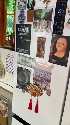 The video shows the interior of a refrigerator and its contents. On the door of the refrigerator, there are various magnets and pictures, including a cutout of Che Guevara, a hand-shaped magnet, a poster for "Kagami," a Beatles Abbey Road album cover, and a rendition of the Mona Lisa. Inside the refrigerator, there are shelves stocked with food items. The top shelf holds a yellow container and a glass jar. The second shelf contains what appears to be produce wrapped in paper and a halved watermelon. The third shelf has a bag of white contents and a large metal bowl. The fourth shelf displays two small bowls, one with red berries and another with a lemon, along with a bunch of yellow objects, possibly bananas, and green vegetables. The bottom drawer contains more food items. The refrigerator door is slightly ajar, revealing a white bottle and a carton of milk on a side shelf. The environment appears to be a kitchen, with a window and plants visible in the background.