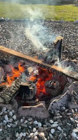 A campfire burns with bright orange flames and heavy white smoke. A wooden board rests across some of the burning wood. The fire is contained within a ring of large stones, surrounded by a bed of smaller grey and white pebbles. Green grass is visible beyond the fire. A car can be faintly seen in the distance. The video appears to be taken outdoors, likely during daylight.