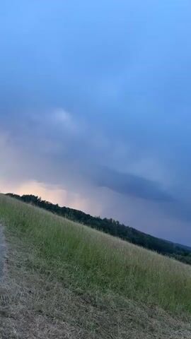 A video shows a landscape at dusk. The sky is a deep blue with dark, ominous clouds that appear to be gathering. In the foreground, a grassy hill slopes downwards and to the right. The grass is a mix of green and dried yellow, indicating a change of season or recent dry weather. The hill is partially covered by a rough texture, possibly dirt or gravel, suggesting a path or uneven terrain. In the background, a dense line of trees forms a dark silhouette against the sky, marking the edge of a forest or wooded area. Beyond the trees, a small town or village is visible, with houses and buildings partially illuminated by the fading light. The overall atmosphere of the video is calm but also foreboding, possibly due to the approaching storm suggested by the clouds.