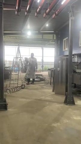 This media file shows an indoor workshop setting, likely a metal fabrication or welding shop, located in Sokury, Russia. The scene is well-lit, possibly during daylight hours, with sunlight filtering in from an open large bay door.

The primary subject is a person, dressed in a grey apron and dark pants, standing near a ladder in the left-center of the frame. They appear to be holding a tool or equipment, possibly related to welding or metalwork. The person is the only human present.

Scattered throughout the workshop are various objects and equipment indicative of metalworking. These include a ladder, coiled hoses, metal beams and structures, large metal cabinets or enclosures, workbenches laden with tools and materials, and what appears to be a large piece of assembled metal equipment in the foreground and middle ground. The overall impression is one of an active or recently active workspace.

The setting features a concrete floor and industrial-style walls, with blue siding visible on the rear wall, punctuated by several windows. Overhead, there are industrial lights and a metal ceiling structure. The presence of large metal machinery and workbenches suggests a focus on heavy fabrication.

The activity taking place appears to be related to metalworking or manufacturing. The person's presence and the surrounding equipment suggest work is in progress or has recently concluded. There is no discernible text within the video. The atmosphere seems utilitarian and industrious.