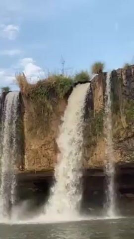 The video shows a low angle view of a waterfall.  The waterfall has four or five separate streams of water flowing over a cliff. The cliff is made of light brown earth and topped with greenery. The sky is blue with light clouds. The water at the bottom of the waterfall is calm.