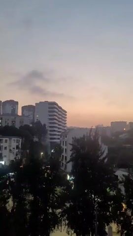 The video shows a cityscape at dusk. In the foreground, there are trees, partially obscuring the view. Beyond the trees are buildings of varying heights and architectural styles, mostly white or light-colored. In the background, the sky transitions from a pale blue at the top to a soft orange hue near the horizon. Some clouds are visible, scattered across the sky. City lights twinkle throughout the scene, illuminating streets and buildings. In the distance, a tower-like structure can be seen atop a hill. The camera appears to be stationary, filming from an elevated position, possibly from a window or balcony.