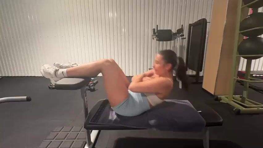 A young woman with dark hair pulled back in a ponytail is performing crunches on a weight bench in a gym. She is wearing a white sports bra and light blue shorts. Her feet are hooked under the padded support at the end of the bench, and her hands are clasped at her chest. The bench is covered with a dark towel. In the background, there is gym equipment, including a wall with corrugated metal siding, a multi-functional weight machine, and a rack of medicine balls. The lighting is artificial, creating a well-lit environment. The woman is engaged in a repetitive motion, contracting her abdominal muscles to lift her upper body towards her knees and then returning to the starting position.