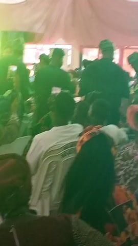 The video shows a crowd of people gathered in an indoor space, possibly a hall or tent, with draping fabric overhead. Many are seated and appear to be observing something in front of them, possibly a performance or ceremony. Some individuals are wearing traditional attire, including hats, and others are using their phones, possibly to record or take pictures. The scene is lively and active, with a sense of anticipation or celebration in the air.