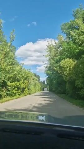 A first-person perspective video shows a car driving down a tree-lined road on a sunny day. The road is asphalt and appears to be two lanes. Lush green trees and foliage line both sides of the road. The sky is blue with scattered white clouds. In the distance, a white structure, possibly a small house or shed, is visible. The camera appears to be mounted inside the car, looking out through the windshield. The sun is casting shadows on the road.