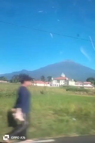The video shows a view from a moving vehicle looking out at a landscape. In the foreground, there is green grass that appears blurred due to the motion. In the midground, there is a building with a light-colored facade, a red-tiled roof, and a dome-like structure. Several other buildings with red roofs are visible to the left and right of the main building. Beyond the buildings, there is a large, conical mountain dominating the background under a clear blue sky. There are some trees and vegetation in the scene, and a utility pole is visible on the right side of the frame. A watermark in the bottom left corner reads "OPPO A76".