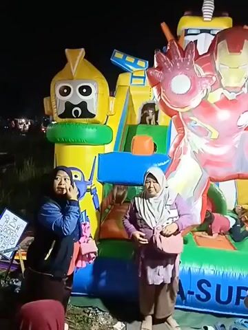 The video captures an outdoor nighttime scene with an inflatable bounce house as the central element. The bounce house is large and colorful, featuring prominent figures of Iron Man and a yellow robot with a black face. Several children are visible inside and on the bounce house, appearing to play. Adults are gathered around the base of the structure, some observing, others seemingly engaged in conversation. One woman on the left side of the frame is wearing a blue jacket and a headscarf, and she is holding her hand to her face. Another woman in the center is wearing a purple jacket and a patterned headscarf, carrying a bag. The ground around the bounce house is dark and appears to be uneven, with some discarded footwear visible. In the background, there are hints of other activities or stalls, suggesting a public event or fair. The lighting is a mix of ambient darkness and the illumination from the bounce house itself and possibly nearby lights.