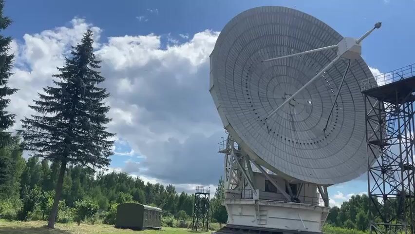 A large, parabolic satellite dish antenna is shown from a low angle, tilted upwards. The antenna is white and mounted on a white structure with a white counterweight visible below. To the right of the antenna, a tall, black metal tower with platforms and railings rises upwards. In the background, a dense line of green trees forms a treeline under a blue sky with scattered white clouds. To the left of the antenna, a tall, dark green evergreen tree stands prominently. A small, olive-green rectangular structure, possibly a shelter or equipment shed, is visible in the grassy field in front of the treeline and to the left of the antenna's base. The ground is covered in dry grass. The sun is shining, casting shadows. There are no people visible in the video.