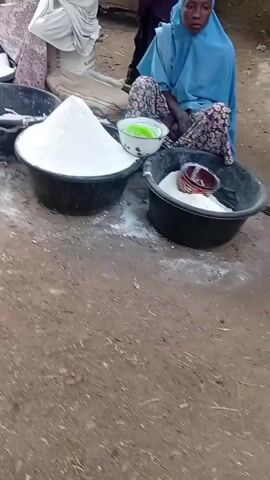An outdoor market scene features three women sitting on dirt ground. In the foreground, two large black plastic basins are filled with mounds of a white powdery substance, likely flour or semolina. One basin contains a small white bowl holding a green liquid, and another small black bowl. A woman wearing a blue head covering and floral pants sits behind these items. To her left, another woman in a white head covering and pink skirt is seated near a similar basin of white powder and several large white sacks. A third woman in an orange head covering is also visible near additional basins of white powder. The background reveals other market activities, including standing individuals, corrugated metal structures, and stalls displaying various goods, notably dried red peppers. The ground is unpaved dirt throughout the visible area.
