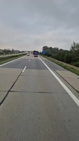A POV shot from a vehicle on a highway shows a cloudy day. The road has multiple lanes and is marked with white lines. Several vehicles, including cars and a truck, are visible on the road ahead. A blue highway sign indicating "Hengersberg 1000 m" is visible on the right side of the road. The surrounding environment includes a grassy median and trees. The road surface appears to have some patched areas.