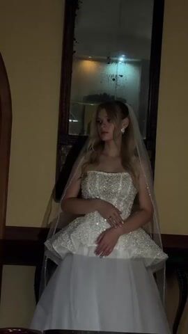 A young woman in a white wedding dress stands in front of a mirror, looking to her right. The dress has a strapless, embellished bodice with a peplum and a full skirt. She is wearing a veil and earrings. The background appears to be an indoor setting with yellow wallpaper and a dark wooden frame around the mirror. The lighting is dim and warm.