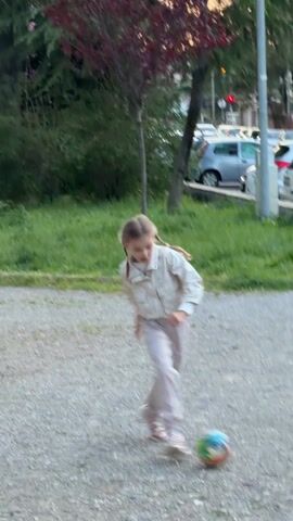 A young girl kicks a colorful soccer ball in an outdoor gravel area near a street with parked cars. The girl is wearing a light-colored jacket, pants, and sneakers. She is fair-skinned and has blonde hair in pigtails. After kicking the ball, she throws her arms out dramatically. A younger boy, wearing a dark blue jacket, runs into the frame toward her. The background includes green grass, trees, and a line of parked cars.