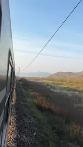 The video shows a view from a moving train. The side of the train is visible, with windows reflecting light. Outside the train, the landscape is a mix of green and brown vegetation, with a hill visible in the distance under a blue sky with scattered clouds. Overhead electrical lines and support structures pass by as the train moves. The overall impression is of a train journey through a rural area on a clear day.