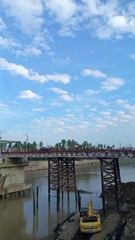The video shows a river with a metal bridge spanning it. The bridge is decorated with red and white flags, and several people are walking across it. The river banks are muddy, and a yellow excavator is visible on a platform near the water. The sky is blue with scattered white clouds.
