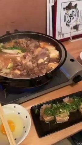 The video shows a simmering pot of sukiyaki on a portable burner, with tofu dishes and a bowl of raw egg nearby on a wooden table. The sukiyaki contains beef, vegetables, and tofu in a broth. The camera zooms in on the sukiyaki, showing the steam rising from the pot. A menu is visible in the background.
