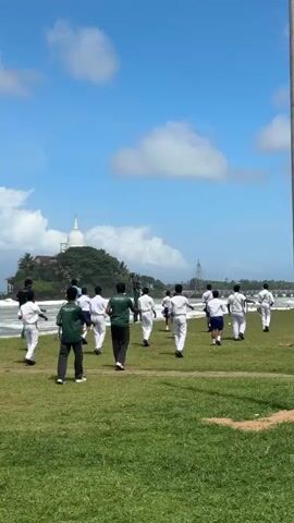 A group of people, mostly young males dressed in white uniforms with some in green shirts, are standing and walking on a grassy field. They are facing towards the ocean where waves are crashing. In the background, on a small island, there is a white stupa or temple with a spire. The sky is blue with scattered white clouds. The overall scene suggests an outdoor activity or training session near the sea.