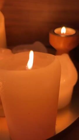 A close-up shot shows several lit candles with warm, glowing flames. The primary candle in the foreground is a thick, white cylinder with a soft glow. In the background, a smaller candle is visible in a wooden holder, and another white candle is out of focus. The lighting is dim, creating an intimate and serene atmosphere, with the warm light casting shadows and highlighting the textures of the candles and their surroundings. The background appears to be a wooden surface, adding to the cozy ambiance.