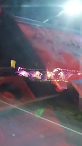The video shows a close-up, shaky view of a structure that appears to be part of an amusement park ride or display. The structure is brightly lit with various colored lights, including red, orange, purple, and green. There is a large graphic on the side of the structure, which is difficult to discern clearly due to the lighting and motion, but appears to depict several white horses in motion against a red and white background. The structure itself is made of metal and has visible bolts and hinges. The overall environment seems to be outdoors at night, with darker areas visible in the background. The camera is in constant motion, moving around the structure and tilting upwards and downwards.