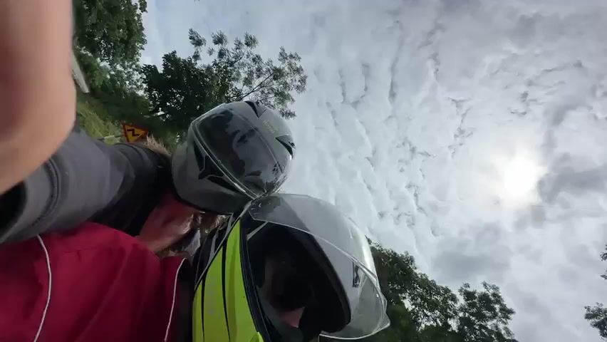 The video shows two people on a motorcycle. The rider is wearing a red jacket and a black helmet. The passenger is wearing a gray hoodie and a black and gold helmet. They are riding on a paved road that curves to the right. The road is bordered by a grassy embankment on the left and trees on the right. The sky is cloudy with patches of blue. A yellow sign is visible on the embankment.