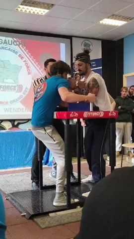 A video shows two men engaged in a contest of arm wrestling. The men are positioned at a table with a red and blue arm-wrestling pad. A referee stands behind them, monitoring the match. The man on the left is wearing a blue t-shirt and gray sweatpants. The man on the right is wearing a white t-shirt and black pants, with a headband holding back his hair. The referee is wearing a striped black and white shirt and dark pants. A crowd of onlookers are in the background, watching the competition. The setting appears to be a community center or similar indoor space, with a projector screen displaying the event's logo and sponsors. The event is titled "FORCE GRAND EST". The man on the right wins, quickly pinning the other man's hand to the pad.