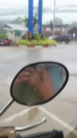 The video shows a view of a gas station during a rainstorm. The camera is positioned at a low angle, possibly from the perspective of someone on a motorcycle, as a side mirror is visible in the foreground with raindrops on it. In the background, a PTT gas station sign is visible, along with utility poles and wires. A pink motorcycle is parked to the left of the gas station, and there is a small white booth or structure in the middle ground. The ground is wet and reflective from the rain. The weather appears overcast and stormy.