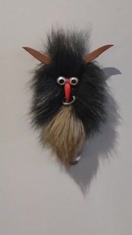 A close-up shot shows a homemade Krampus-like ornament hanging against a plain white wall. The ornament is primarily composed of black faux fur, with a tan-colored faux fur beard. It has large, googly eyes and a red, cone-shaped nose. A silver hoop is attached to the ornament, likely as a nose ring. The horns are made of a flat, brown material, possibly leather or felt. The shadow of the ornament is cast on the wall.