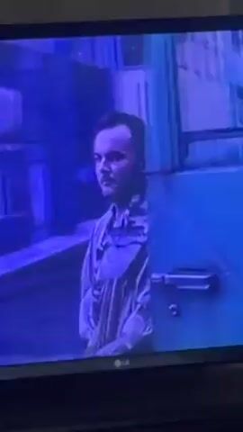A man with dark hair pulled back in a short ponytail is seen on a television screen. He's wearing a light-colored button-up shirt, possibly open, and a translucent jacket. He stands in what appears to be an urban setting, with buildings and possibly a parked car visible in the background. The overall scene has a blueish tint due to the screen's color balance. The television has an LG logo in the lower-center.