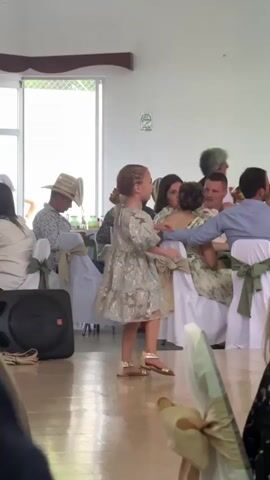 The scene is indoors, seemingly at a wedding reception or similar event. A young girl, dressed in a short, puffy, silver-patterned dress and gold sandals, is standing on a light-colored dance floor or open space. She is turned towards a table where several people are seated. Some of the seated people are interacting and talking, while others are looking down. One person is wearing a straw hat. The table is decorated with white tablecloths and chairs tied with green ribbons. To the left, a black speaker stands on the floor. A large window is visible in the background. The room is well-lit. A part of a draped fabric is visible in the foreground. The camera appears to be handheld and slightly shaky.