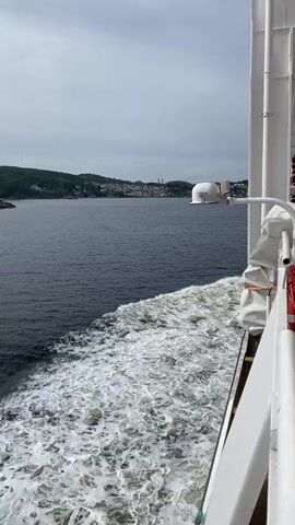 A video from a ship shows a wake being left behind in the dark water of a bay or fjord. The shore is lined with trees and buildings, and the sky is overcast. There is a white dome on a pole on the right side of the frame.