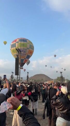 A crowd of people stand in a large open area with a view of pyramids in the distance. Hot air balloons of various colors are floating in the sky, one large one is marked "Sky Balloons MEXICO". Many people are holding up cell phones, presumably taking pictures or video of the scene. There are large speakers visible on towers. The scene is in daylight with a blue sky and scattered clouds.