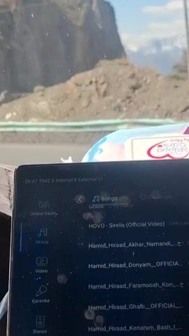 The video, taken from inside a car, shows the vehicle navigating a winding mountain road. The driver's hands are briefly visible on the steering wheel. The dashboard is partially visible, including a screen displaying music information and a navigation system. The car's altitude is approximately 2545 meters, as indicated on the screen. The weather appears sunny, with light shining through the windshield. The foreground consists of the interior of the car, including the steering wheel and dashboard, while the background displays the winding road and the surrounding mountainous landscape. There are a few structures visible by the side of the road.