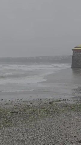 This media file depicts a choppy sea with waves crashing onto a beach in Tramore, Ireland. The weather appears overcast and possibly stormy, with a grey sky and mist obscuring the distant coastline. A stone building with a yellow roof is visible on the right side of the frame, close to the water's edge. The beach itself is covered in pebbles and some scattered seaweed. The waves are white-capped and energetic, suggesting a strong wind. The overall mood is one of wild, untamed nature, with the building serving as a lone structure against the elements. There is no visible text. The time of day is difficult to ascertain due to the overcast conditions, but it appears to be daytime.