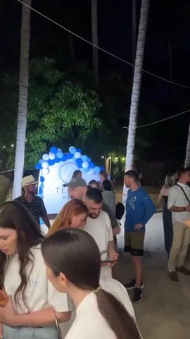 The video showcases an outdoor event at night. The setting appears to be near a beach, identifiable by palm trees. Many people, dressed in casual attire, can be seen mingling. In the background, there's a stage structure with lighting equipment and a screen, potentially for presentations or performances. Tables and chairs are scattered around, suggesting a seated event or lounge area.  A photographer with a reflector is visible, indicating documentation or photography of the event is taking place. The presence of balloons and decorations points toward a celebration or special occasion. People are seen in groups, talking and observing the activity.