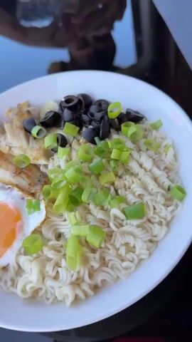 A close-up shot shows a bowl of ramen noodles topped with a fried egg, fish strips, sliced black olives, and chopped green onions. To the left of the bowl, a small white cup contains a white liquid, possibly milk. In the background, another bowl filled with a salad-like mixture is visible. The scene appears to be set outdoors, as indicated by the sunlight and the presence of a wooden railing and potted plants. The camera then pans to reveal a wider view of a patio or deck area. A person is seated at a table, partially visible, engaged in eating. Beyond the table, a wooden fence and a grassy landscape with trees and houses are visible under a clear blue sky. The overall impression is of a casual outdoor meal.