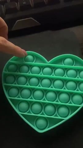 A close-up shot shows a person's hand interacting with a teal-colored, heart-shaped pop-it fidget toy. The toy is placed on a dark, textured surface, possibly a desk or mousepad, with the edge of a black keyboard visible in the upper background. The person's fingers press down on the silicone bubbles of the toy, creating a satisfying popping sound and sensation. The video captures the action from a slightly elevated angle, focusing on the toy and the hand's interaction with it. The lighting appears to be dim, with the main light source illuminating the toy from above.