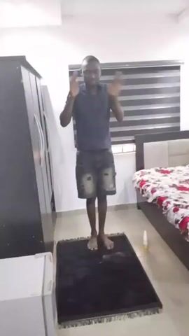 A man is in a bedroom. The room has a window with horizontal blinds, a wardrobe, a bed with a white and red floral patterned comforter, and a small refrigerator. The man is standing on a black rug. He is wearing a dark blue shirt and distressed denim shorts. He brings his hands to the side of his head then he lowers himself to the rug and assumes a kneeling, then a prostrate position.