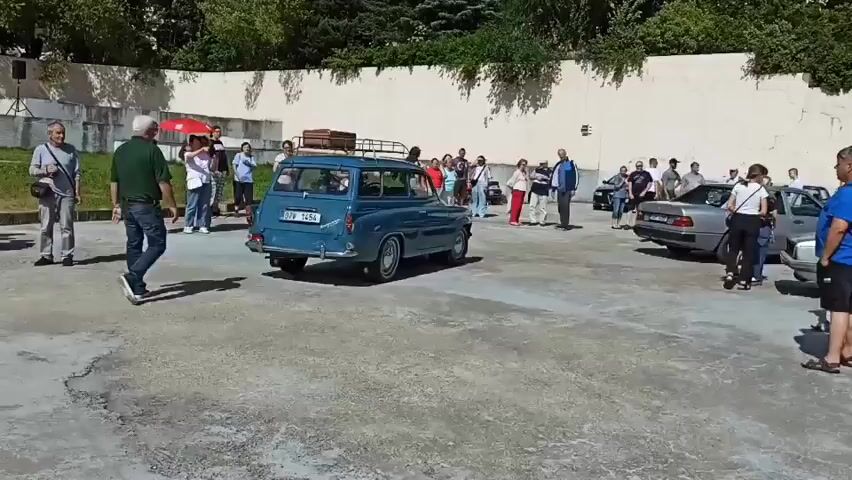 The video shows an outdoor gathering with several vintage cars and people. In the foreground, a bright red vintage car is parked, partially blocking the view of the scene behind it. The car is a four-door sedan with chrome accents and white-rimmed wheels. A person is visible in the driver's seat. To the left and center of the frame, a blue vintage station wagon with a luggage rack on top is moving away from the camera, turning to the right. Several people are standing around the blue station wagon, some observing it. In the background, more people are scattered throughout the paved area, and a long, light-colored wall runs along the back of the scene, with trees and foliage visible above it. There are also other vehicles and some white fencing in the background. The overall impression is of a car show or a gathering of classic car enthusiasts. The lighting suggests it is daytime and sunny.