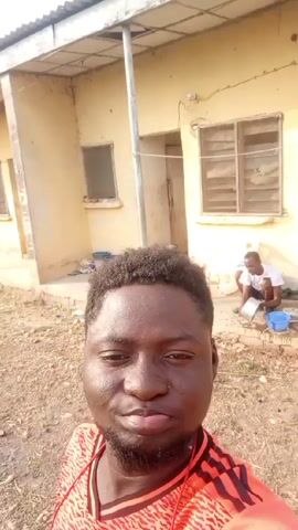The video shows a dark-skinned man in an orange and black shirt holding the camera up to record himself. He sticks out his tongue and smiles.  Behind him is a pale yellow building with an exterior doorway and a window. There is a man washing dishes in the background. The ground appears to be dry and dusty, with sparse vegetation in the distance. The man raises his arm in the air in the last frame.
