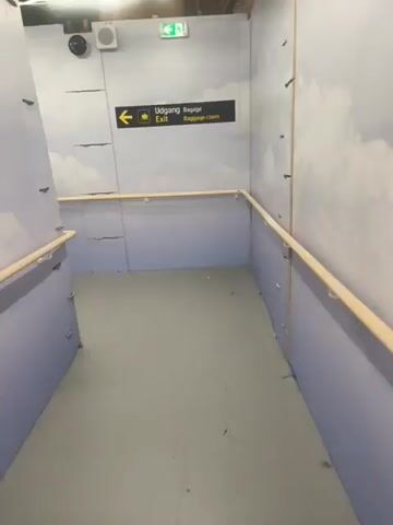 The video shows a hallway with temporary construction-like walls. The walls are light gray with a patterned design resembling clouds. A wooden handrail runs along the lower half of the walls on both sides, supported by metal brackets. The floor is a light gray, smooth surface.

On the left wall, a sign indicates directions. The sign is black with yellow text and symbols. It reads "Udgang Baggage Exit Baggage claim" with an arrow pointing left and a graphic of a suitcase. Above the sign, there's a security camera and a white speaker.

Further down the hallway, the walls are adorned with large text. The visible text on the left wall reads "UPGRADING WE ARE BUILDING A MUCH BETTER AIRPORT". The text on the right wall is partially visible and reads "WE ARE BUILDING A MUCH BETTER AIRPORT".

Two people are walking away from the camera down the hallway. They are walking towards a light source at the far end. The lighting suggests it is daytime, but the indoor setting makes it difficult to determine the exact time of day or weather. The overall impression is that of a temporary walkway, likely within an airport undergoing renovations, given the text on the walls and the "Baggage claim" sign. The environment appears functional and utilitarian, with the cloud pattern adding a touch of aesthetic, albeit in a temporary construction context.