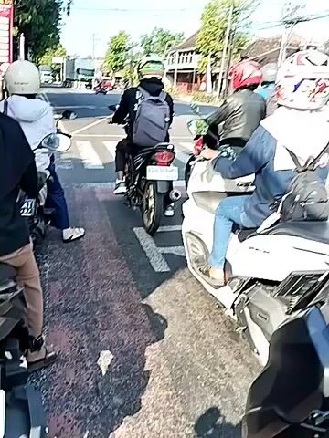 The video shows a street scene in an Asian country with several people on motorcycles waiting at a traffic light.  The motorcycles are a mix of models.  Many of the riders are wearing helmets. Some riders are wearing backpacks. There are power lines overhead and buildings and trees on either side of the road.  A three-wheeled vehicle with some kind of machinery on it is also waiting at the intersection.  When the light changes, the motorcycles accelerate and proceed through the intersection.