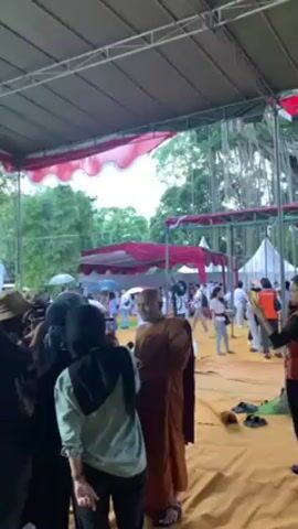 Here is a concise description of the video:

The video shows a Buddhist monk in traditional brown robes surrounded by people, most of whom are taking photos or videos of him with their phones. The scene is outdoors, under a large temporary shelter with a red and white scalloped awning. In the background, there are other tents, trees, and people, some holding umbrellas. One person in an orange vest appears to be an event staff member. The monk seems to be the focal point of attention, and there is a general air of activity and engagement around him.