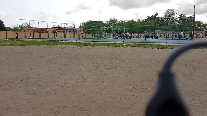The video shows a tennis court from behind a sandy area. There are people standing and sitting around the tennis court, some in what appear to be school uniforms. A net divides the tennis court in half. The court is surrounded by a fence, trees and buildings are visible in the background. The sky is cloudy.