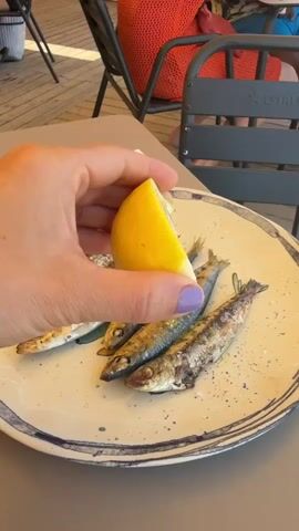 A hand with purple nail polish squeezes a lemon wedge over a plate of cooked sardines. The plate has a blue and white rim. The sardines are arranged in two rows. The background shows a table, chairs, and a person wearing a red top. The setting appears to be an outdoor dining area.