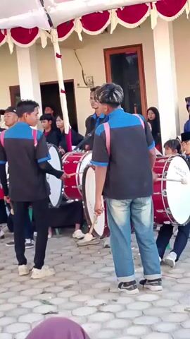 A group of young people in matching blue and black uniforms with pink straps are playing bass drums. Two individuals in the foreground are actively drumming, one on the left and one on the right, both facing away from the camera. The person on the left is holding a white drumstick, while the person on the right is holding a drumstick with a white tip. The drums are large and red with white drumheads. Other members of the group, also in uniform, are visible behind them, some holding drums. The background shows a building with white pillars and a red and white draped canopy overhead. Several spectators, including women and children, are observing the performance. The ground appears to be paved with interlocking stones. The overall scene suggests an outdoor event or performance.