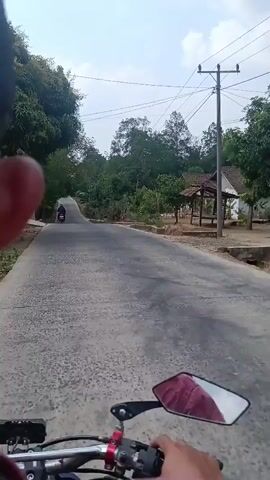 A first-person perspective video shows a motorcycle rider's view as they travel down a paved road. In the distance, another motorcycle is visible traveling in the same direction. The road is flanked by trees and vegetation on both sides. There are also some small structures or houses visible in the background, suggesting a rural or semi-rural setting. The sky is overcast, indicating a cloudy day. The rider's hand and part of the motorcycle's handlebar and mirror are visible in the foreground, confirming the viewpoint.